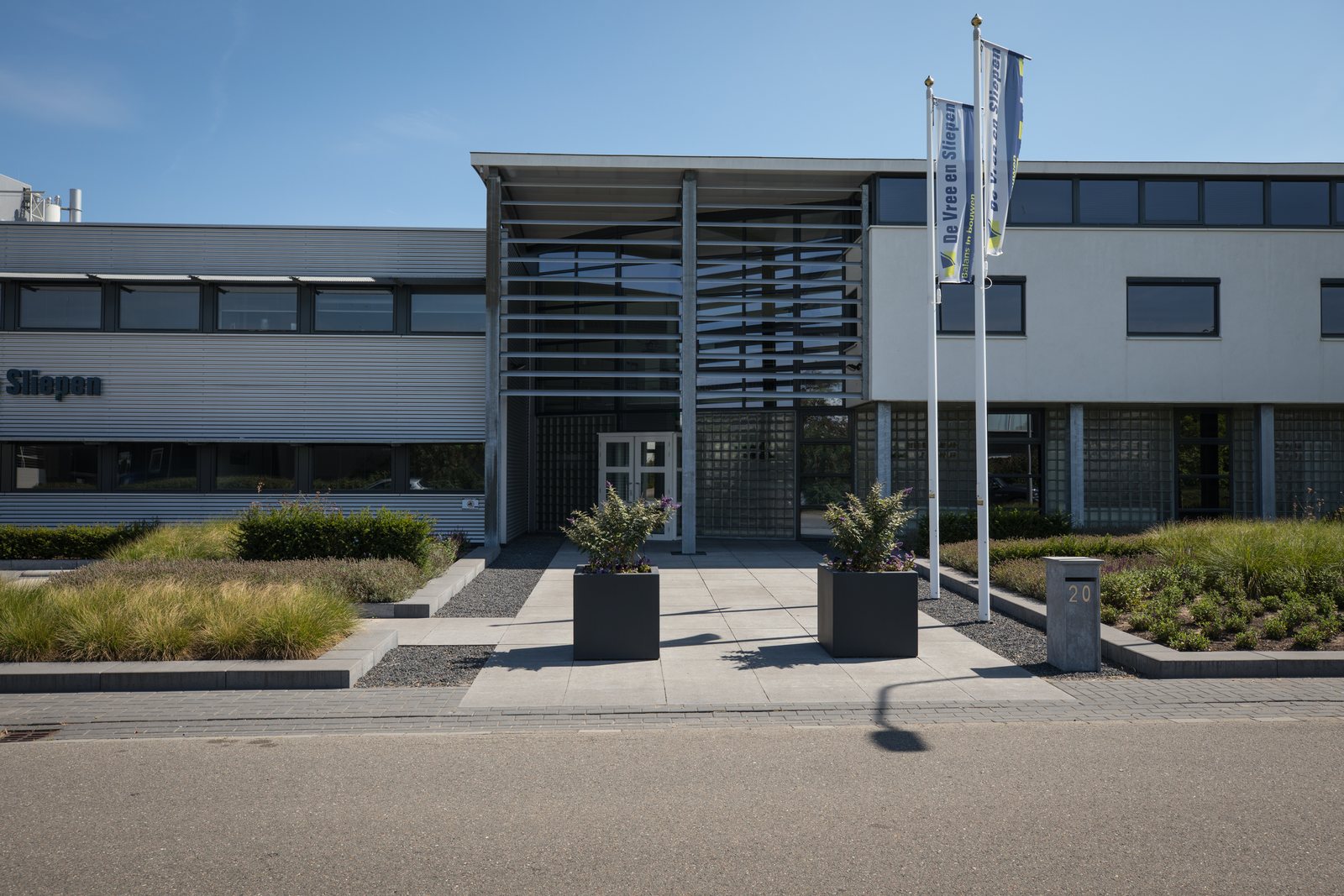 Kantoorpand met bestraat voorterrein, plantvakken met siergrassen en zwarte plantenbakken bij de entree